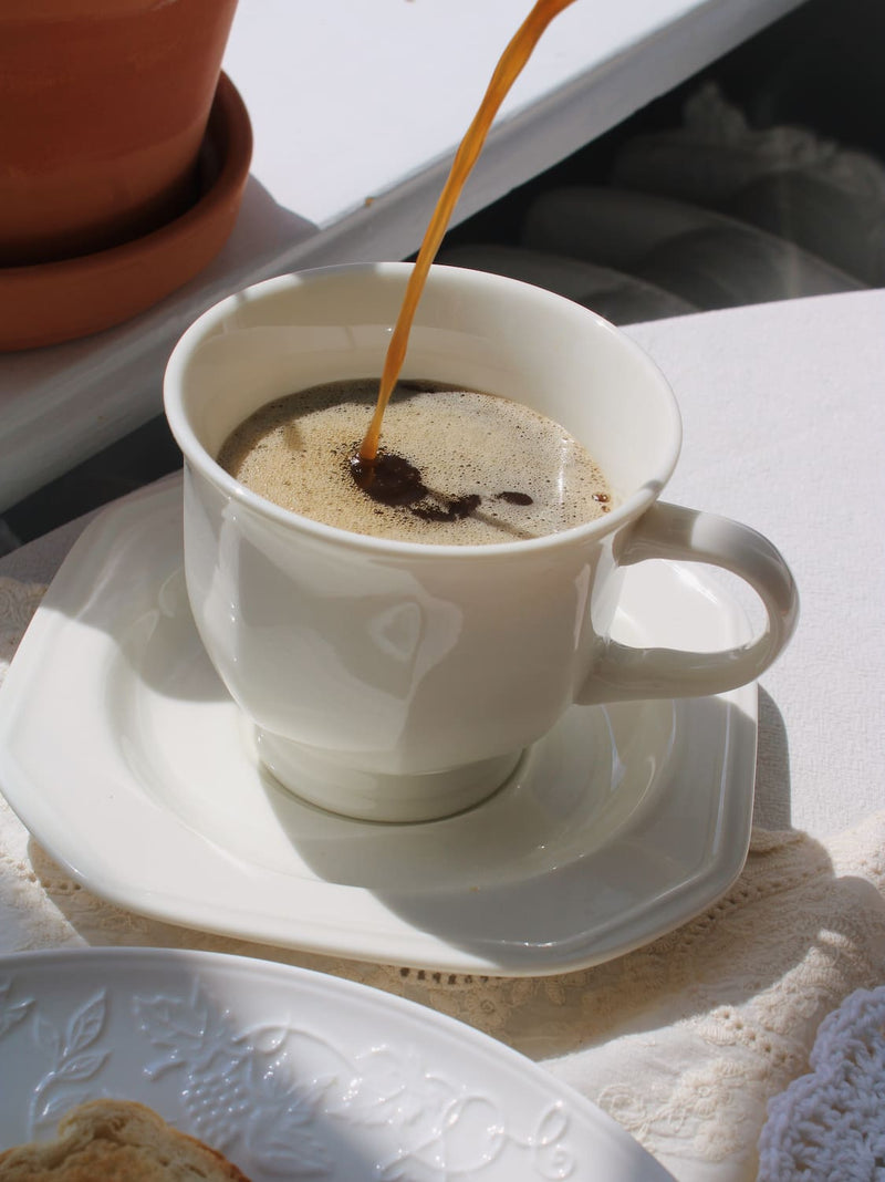 A white ceramic coffee cup on a matching saucer, with coffee being poured into it. The cup and saucer are placed on a white tablecloth, with a terracotta pot in the background and a plate partially visible in the foreground.