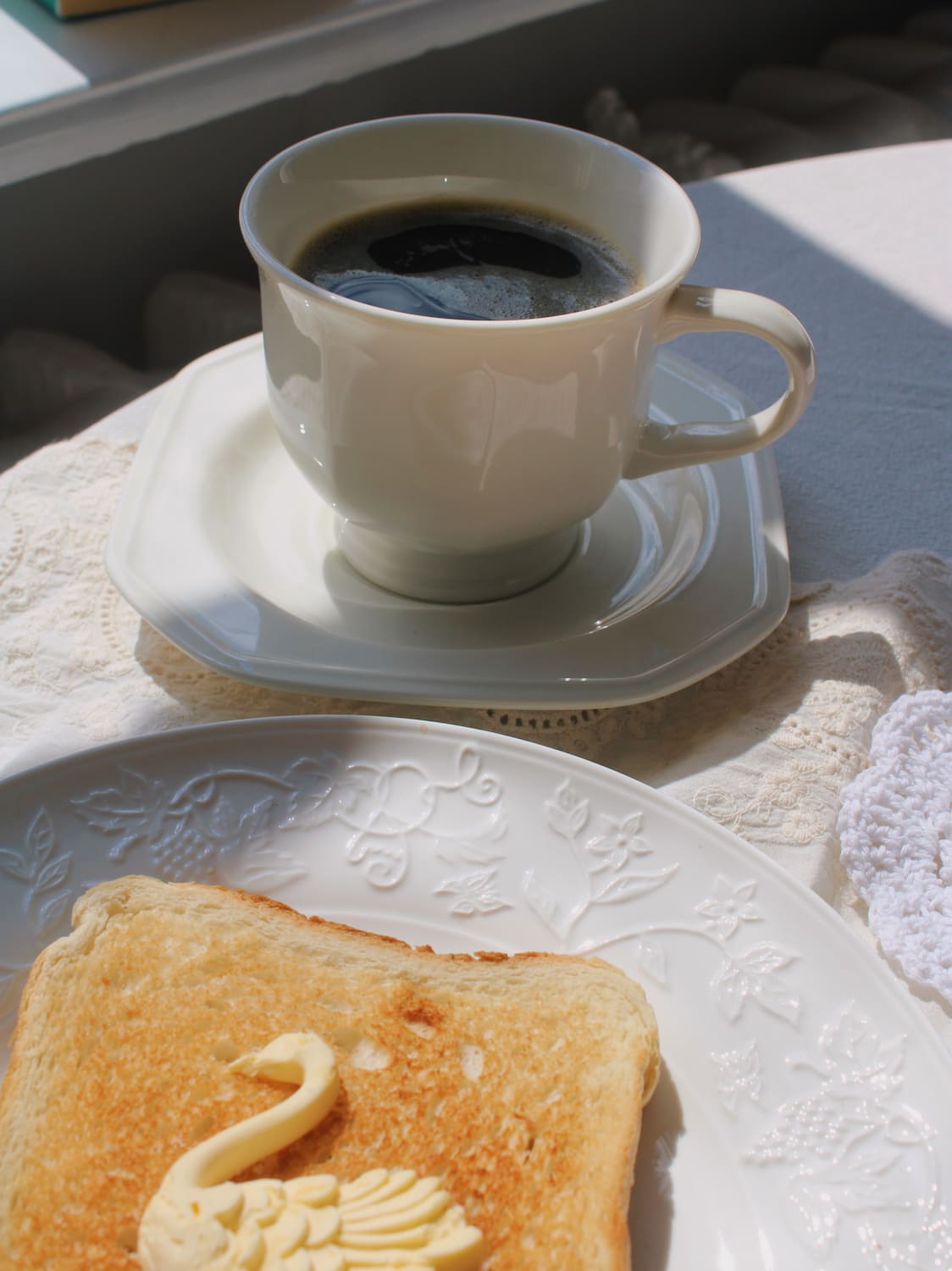 A white ceramic cup of black coffee on a matching saucer, placed on a lace doily next to a plate of toast with a swan-shaped butter decoration.