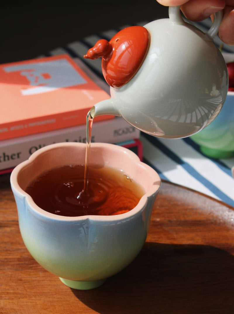 A hand pours amber tea from a glossy white teapot with a reddish-orange lid into a pastel gradient cup with a pink rim, set on a wooden table with books in the background.