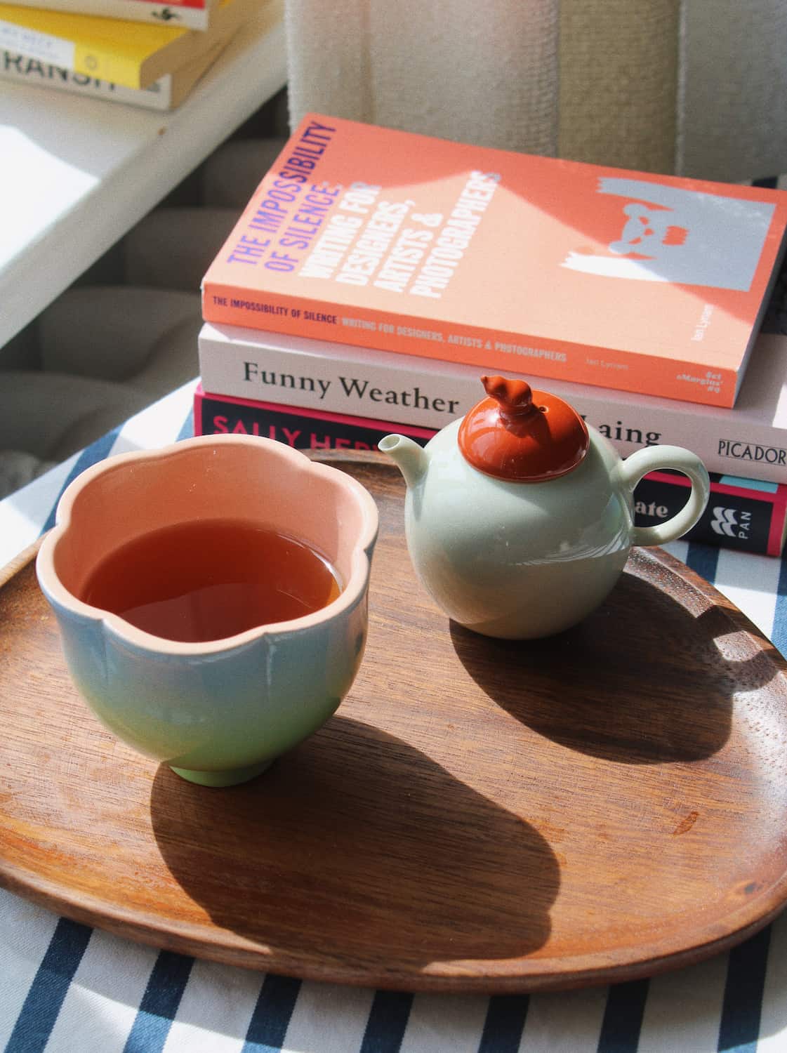 A ceramic tea set with a pale green body and a terracotta-colored lid, alongside a matching flower-shaped cup filled with tea, arranged on a wooden tray with stacked books in the background.
