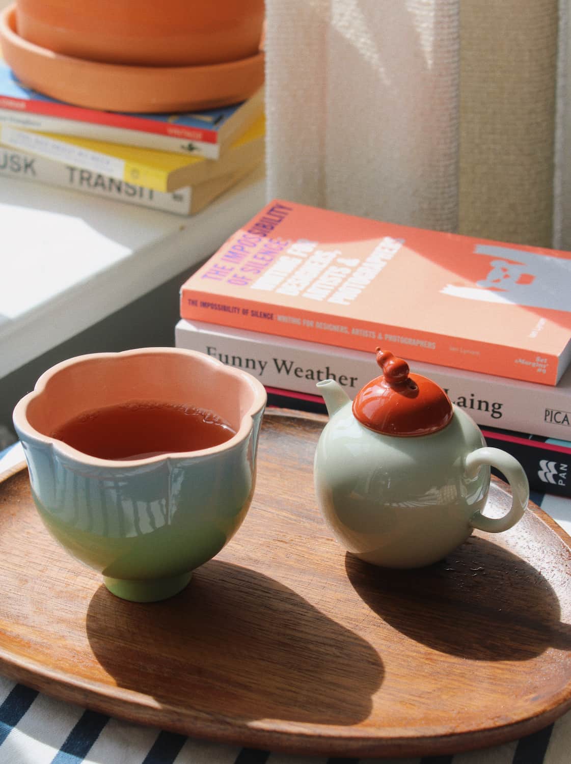 A ceramic tea set with a pale green body and a terracotta-colored lid, alongside a matching flower-shaped cup filled with tea, arranged on a wooden tray next to stacked books in a sunlit indoor setting.