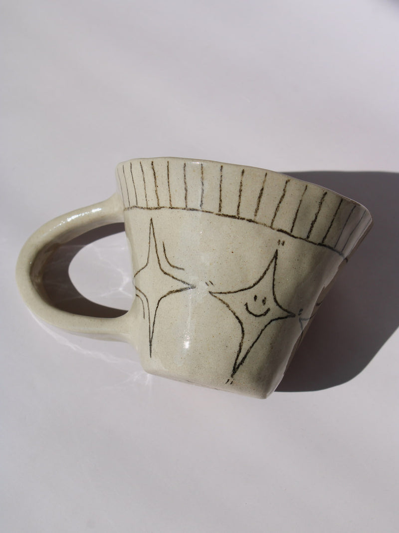 Astral Ceramic Mug in speckled cream with hand-etched four-point star and smiley face motifs and a stripe rim detail, on a soft white background.