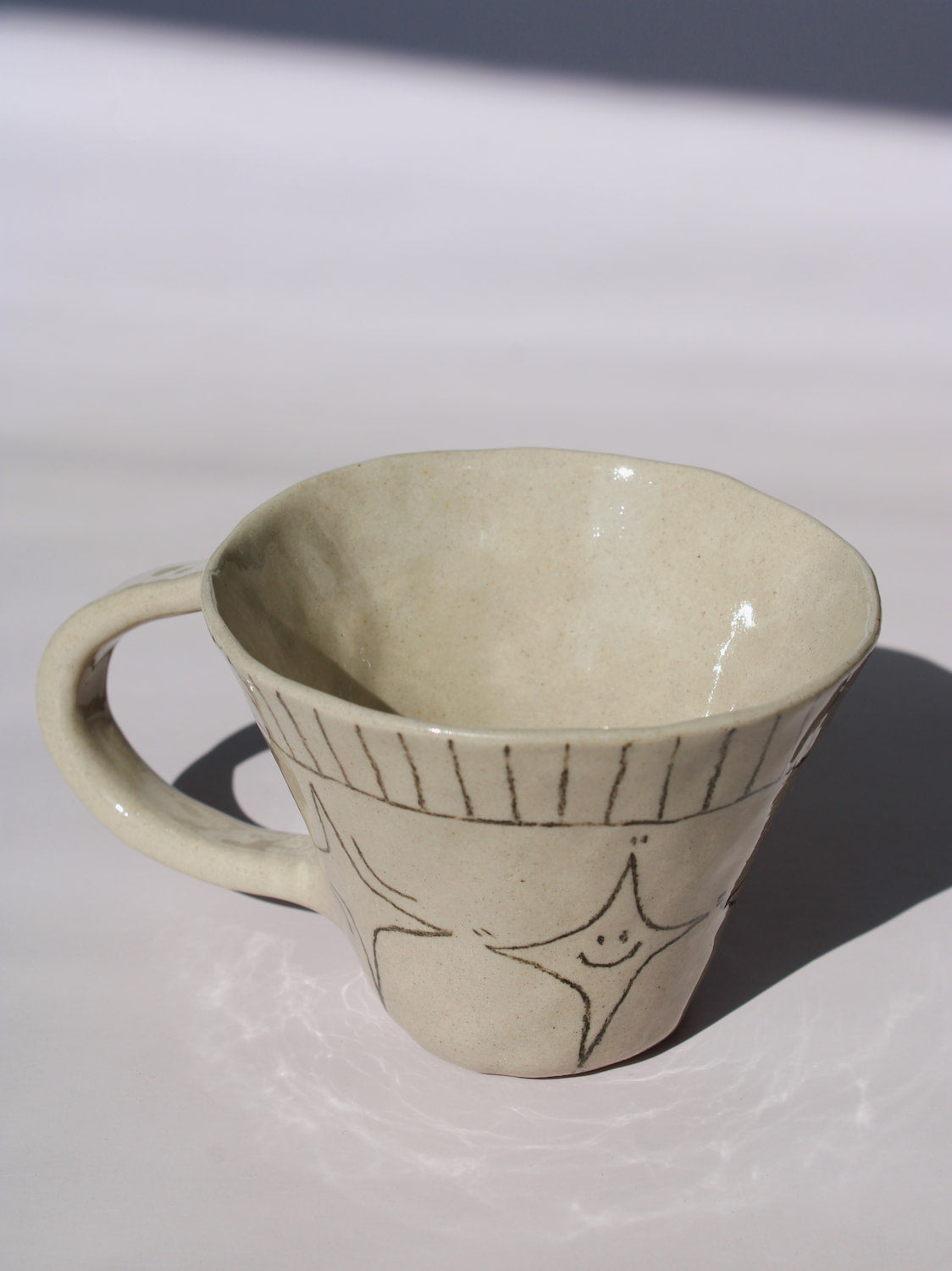 Astral Ceramic Mug in speckled cream with hand-etched four-point star and smiley face motifs and a stripe rim detail, on a soft white background.
