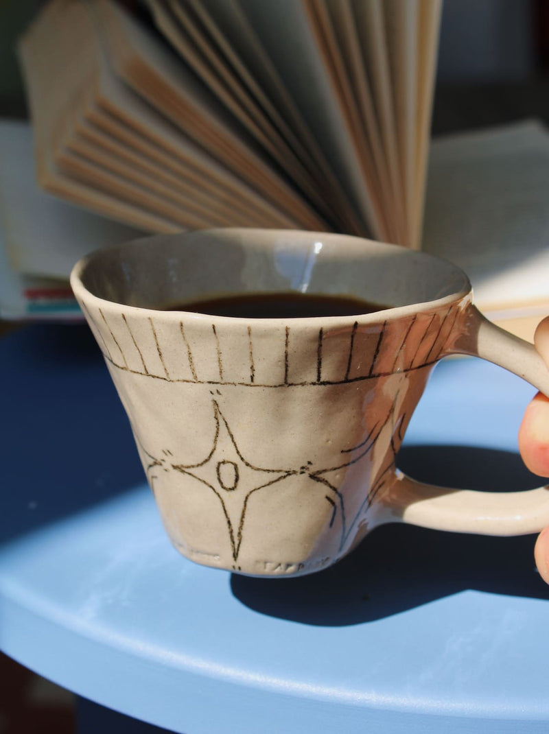 Astral Ceramic Mug in speckled cream with hand-etched star and stripe detail, held in hand filled with a latte, with a blurred indoor background in natural light.