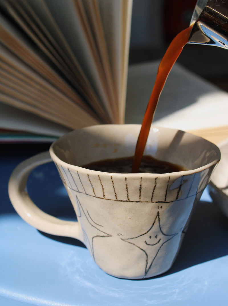 Astral Ceramic Mug in speckled cream with hand-etched star and stripe detail, held in hand filled with a latte, with a blurred indoor background in natural light.