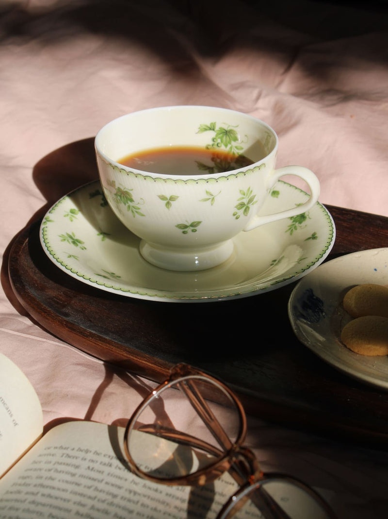 A white ceramic teacup and saucer with a green floral pattern, filled with tea, set on a dark wooden tray. The scene includes an open book, eyeglasses, and a small plate of cookies on a pink fabric background.