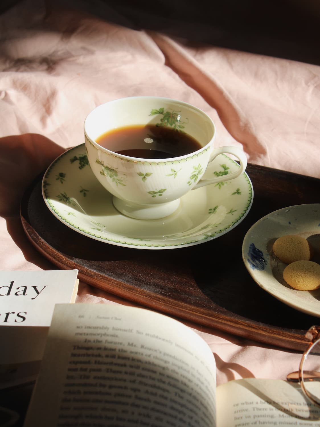 A white ceramic teacup and saucer with a green floral pattern, filled with black coffee, set on a dark wooden tray. The scene is styled with an open book, a small plate of cookies, and soft pink fabric in warm, dappled sunlight.