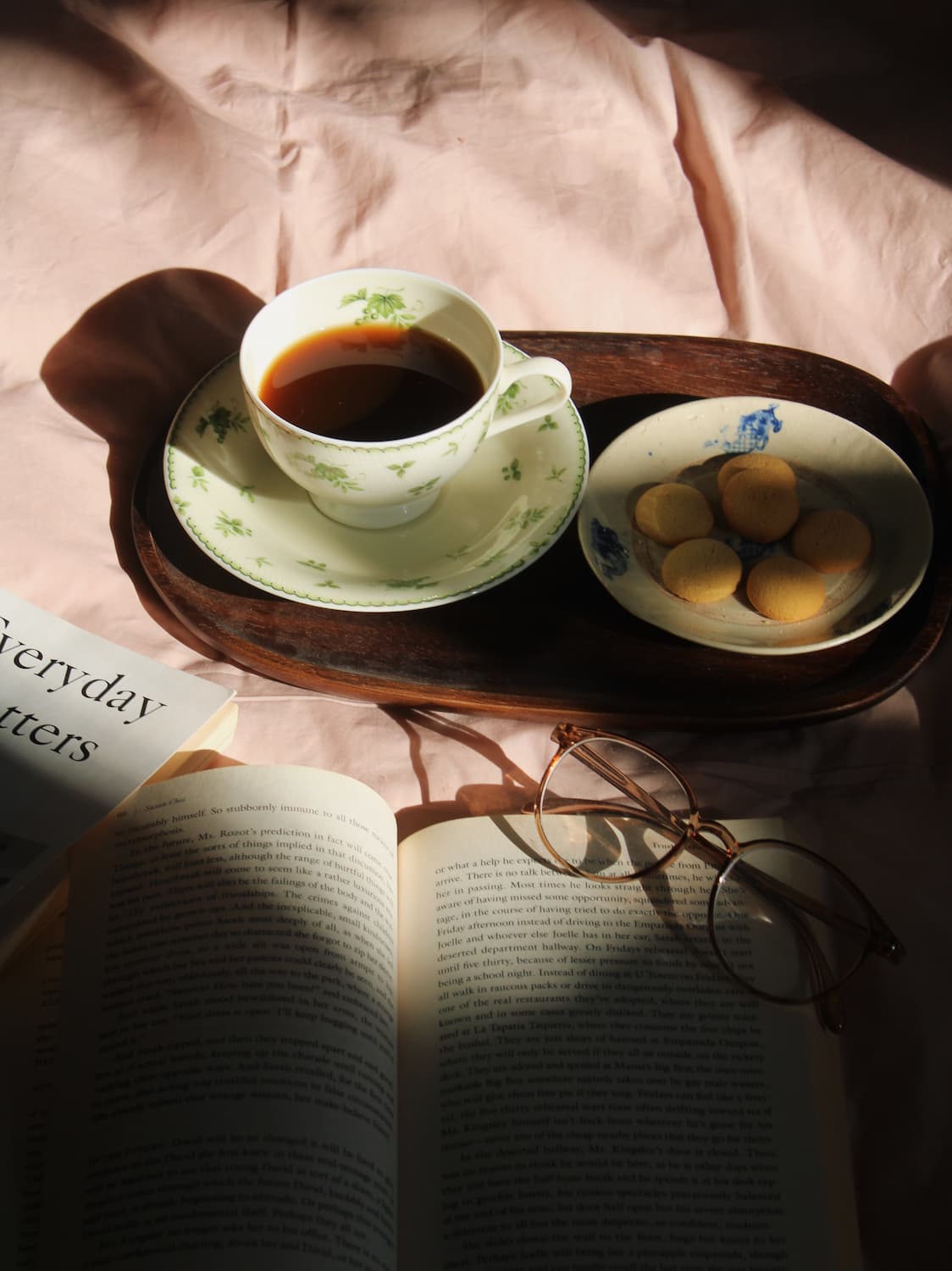 A styled still life with a white floral teacup of black coffee on a saucer, a small plate of cookies, an open book, eyeglasses, and a wooden tray on a pink fabric surface in dramatic sunlight and shadow.