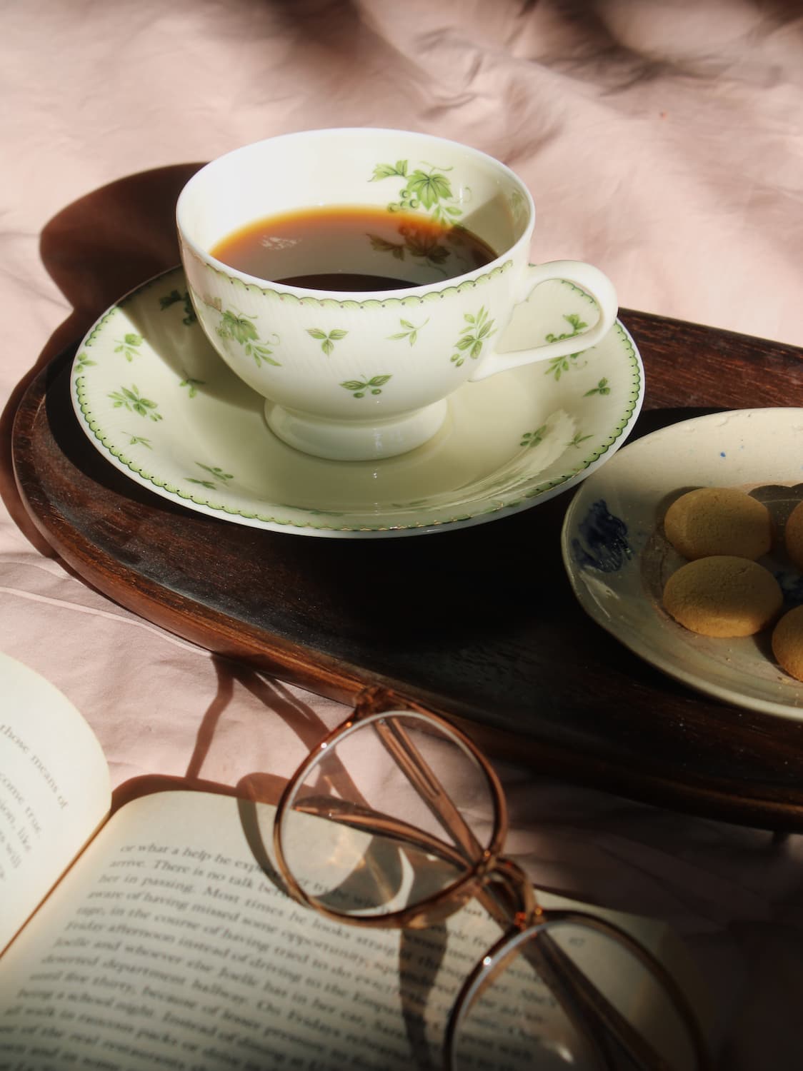 A white ceramic teacup and saucer with a green floral pattern, filled with black coffee, set on a dark wooden tray. An open book with printed text and a pair of round metal eyeglasses rest in the foreground, with a small plate of cookies on the right.