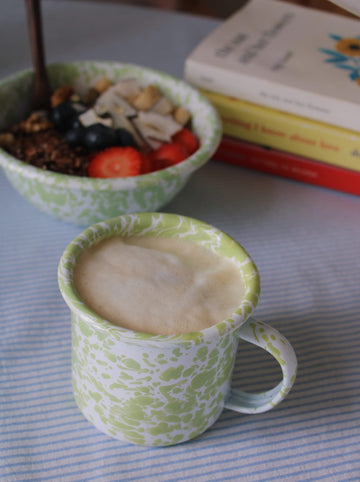 Green mug with a hot beverage on a table with a bowl of fruit and a book in the background.