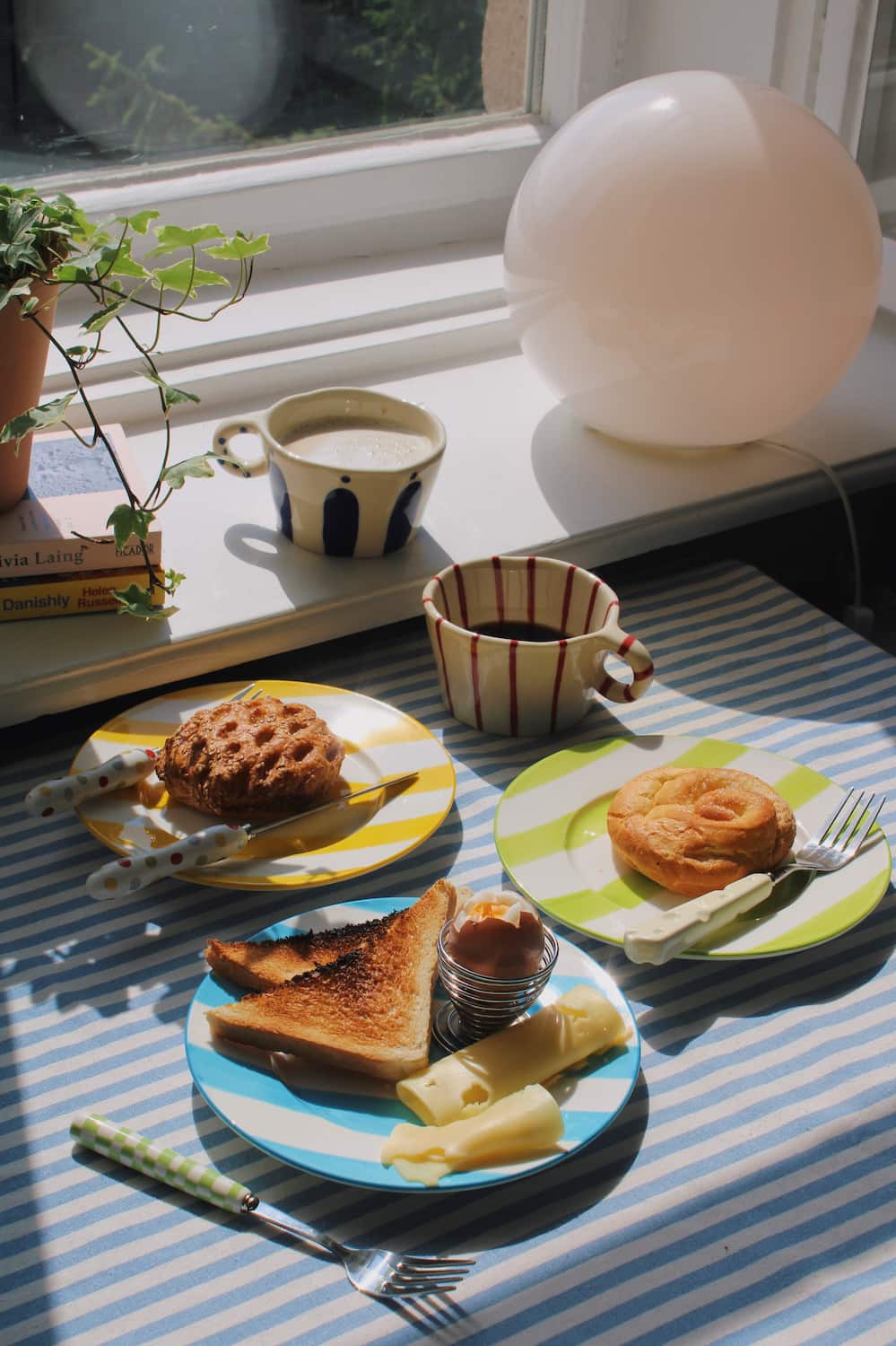 Arlo Ceramic Mug and Enzo Ceramic Mug alongside the Etta Ceramic Plate, Olia Ceramic Plate and Mirel Ceramic Plate styled with a full breakfast spread of pastries, toast, soft boiled egg and cheese on a blue striped surface, with ivy, stacked books and a white globe lamp by the window.