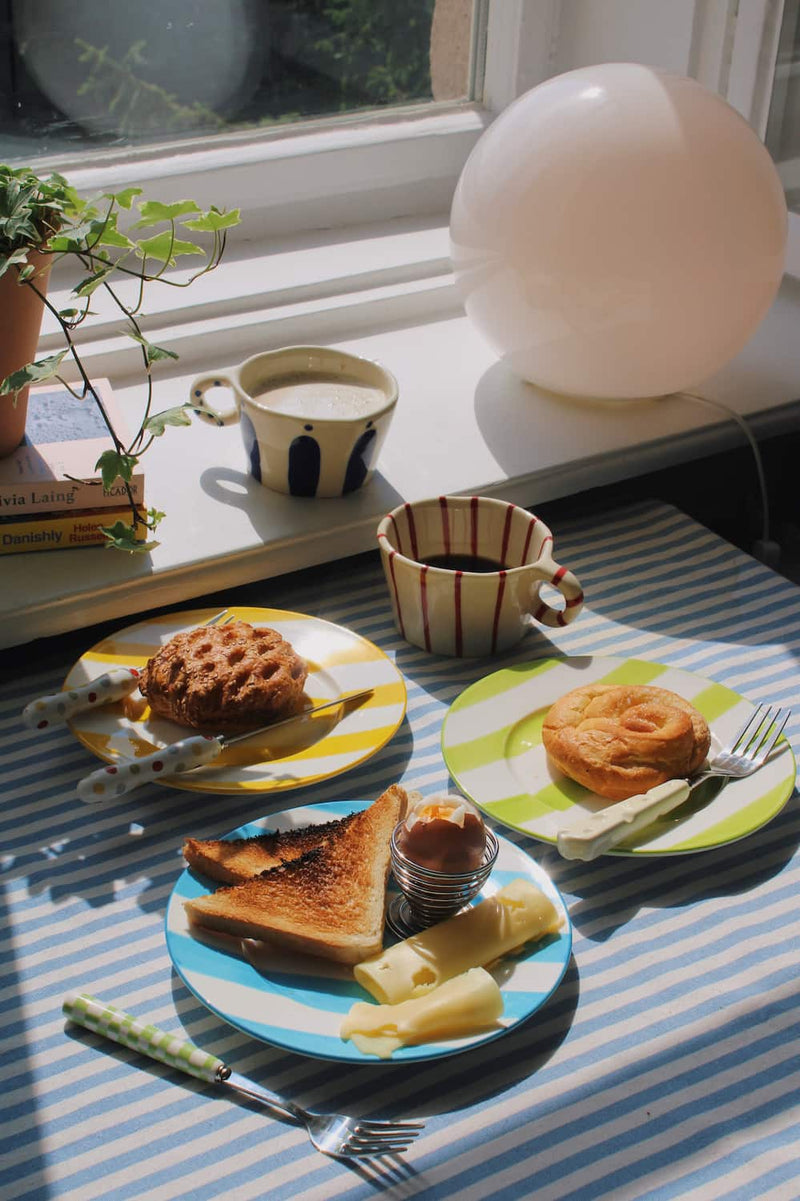 Arlo Ceramic Mug and Enzo Ceramic Mug alongside the Etta Ceramic Plate, Olia Ceramic Plate and Mirel Ceramic Plate styled with a full breakfast spread of pastries, toast, soft boiled egg and cheese on a blue striped surface, with ivy, stacked books and a white globe lamp by the window.