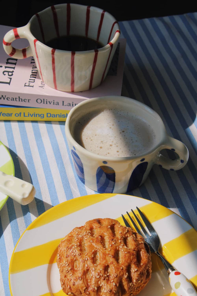 Arlo Ceramic Mug and Enzo Ceramic Mug styled together with a sesame pastry on the Etta Ceramic Plate and the Confetti Cutlery Set, on a blue striped surface with stacked books in the background.