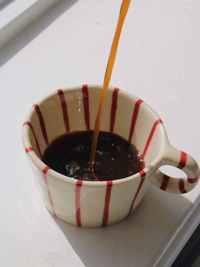 Arlo Ceramic Mug in cream with red vertical stripe pattern, with coffee being poured in, on a white windowsill in natural sunlight.