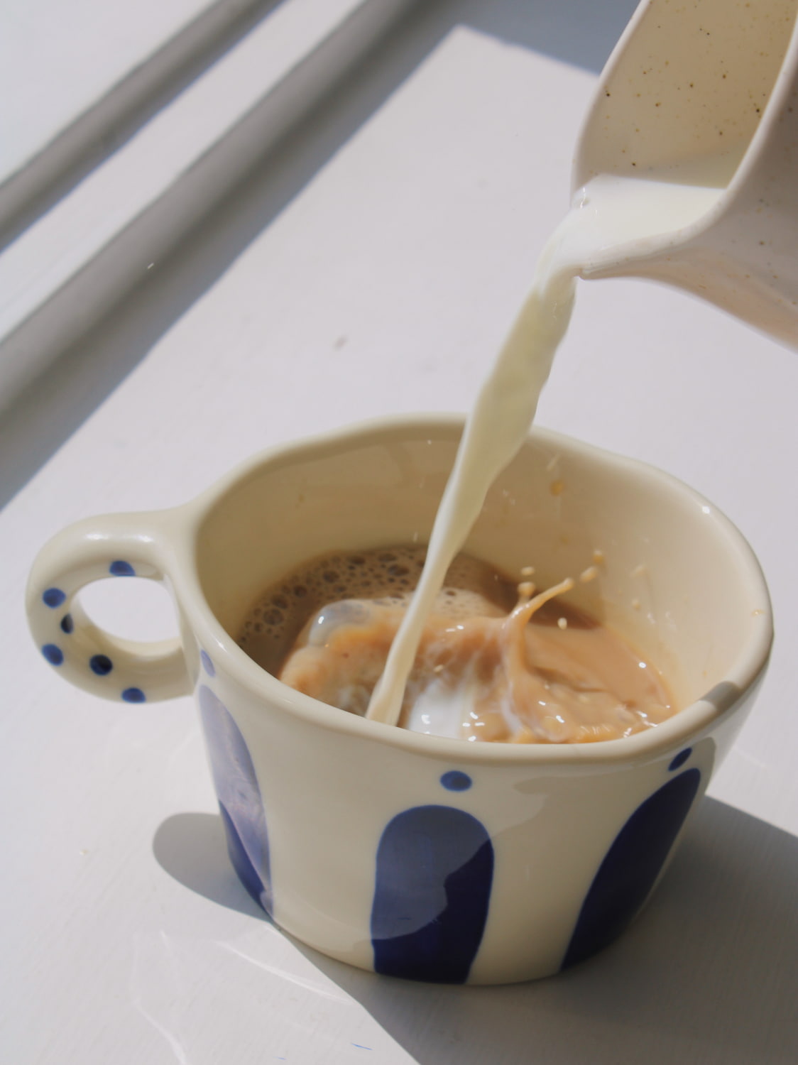 Enzo Ceramic Mug in cream with blue arch and dot pattern, with milk being poured into coffee, on a white windowsill in natural sunlight.