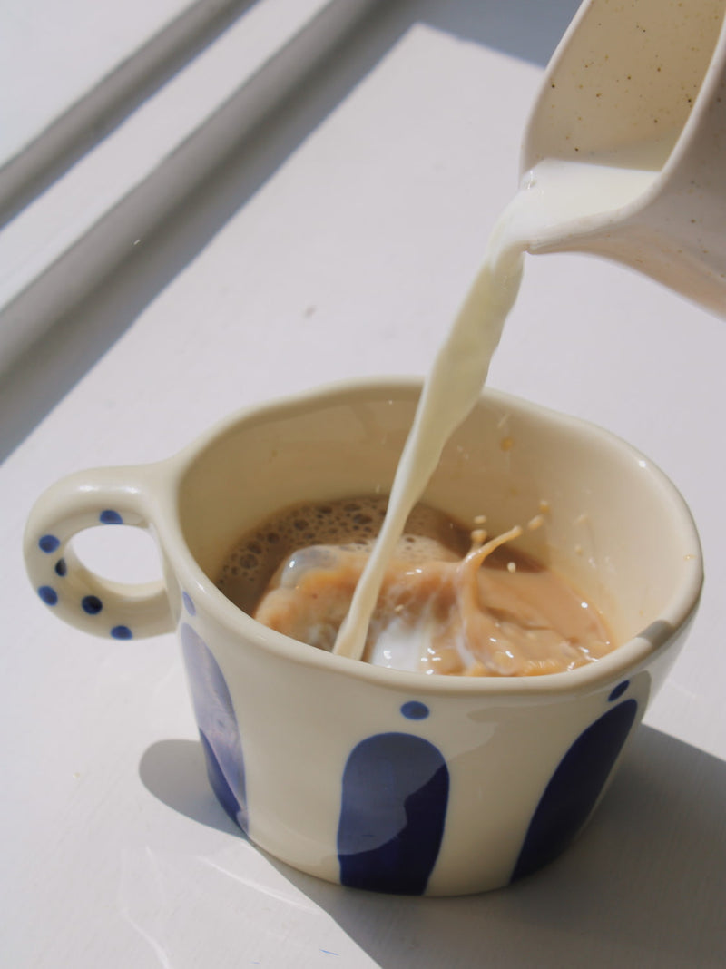 Enzo Ceramic Mug in cream with blue arch and dot pattern, with milk being poured into coffee, on a white windowsill in natural sunlight.
