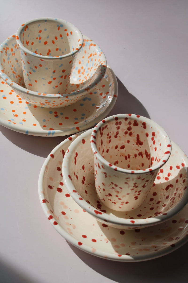 Naro Ceramic Set and Bera Ceramic Set displayed together, showing two matching cup, bowl, and plate combinations in cream — one with orange and blue speckled dot pattern and one with red speckled dot pattern — on a soft pink background.