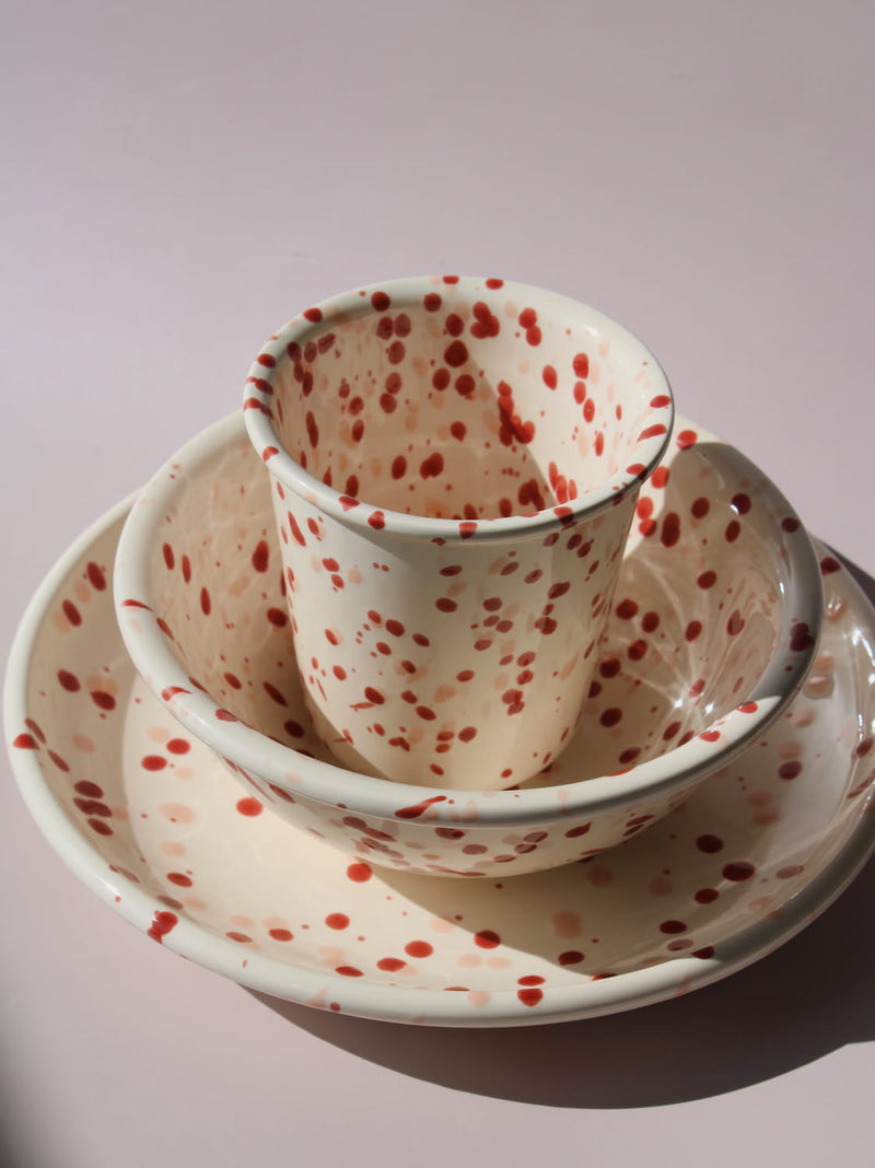 Bera Ceramic Cup, Bowl, and Plate in cream with red speckled dot pattern, on a soft pink background.