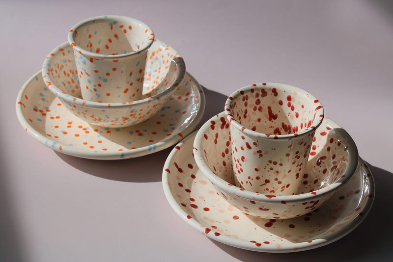 Naro Ceramic Set and Bera Ceramic Set displayed together, showing two matching cup, bowl, and plate combinations in cream — one with orange and blue speckled dot pattern and one with red speckled dot pattern — on a soft pink background.