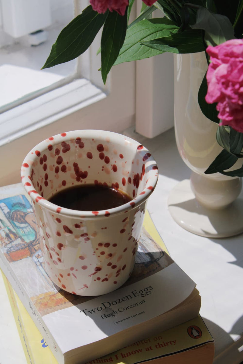 Bera Ceramic Cup in cream with red speckled dot pattern, filled with black coffee and resting on a stack of books by a sunny window, with a white vase of pink peonies in the background.