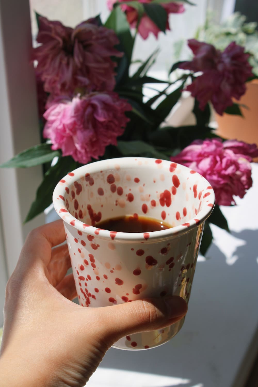 Bera Ceramic Cup in cream with red speckled dot pattern, filled with black coffee and resting on a stack of books by a sunny window, with a white vase of pink peonies in the background.
