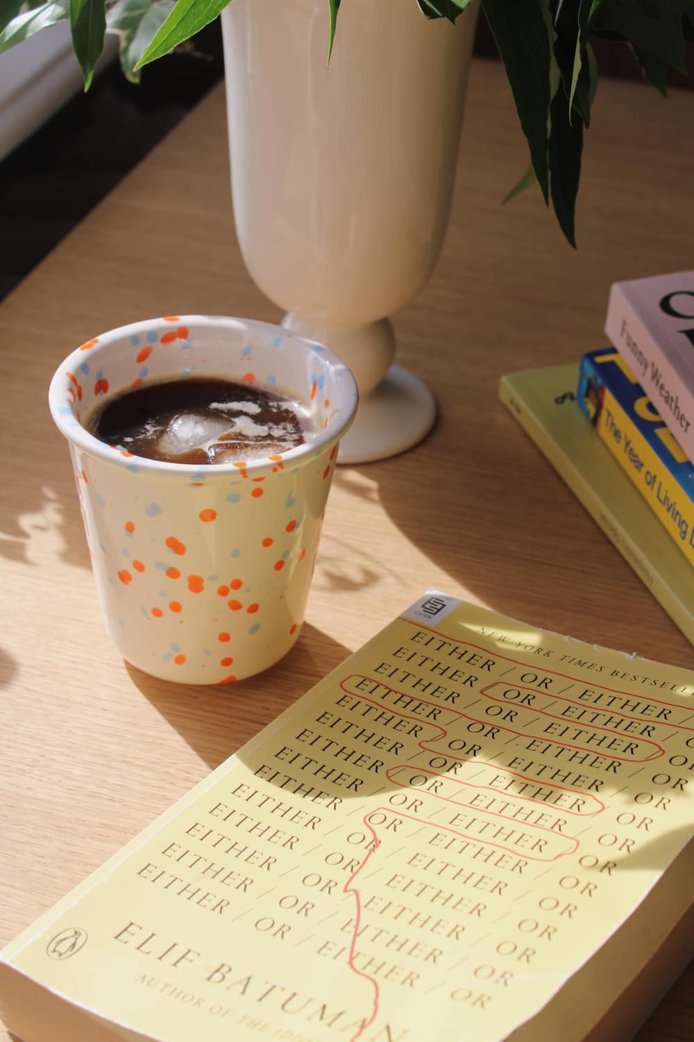 Naro Ceramic Cup in cream with orange and blue speckled dot pattern, filled with iced black coffee on a wooden surface with a book and white vase in natural sunlight.
