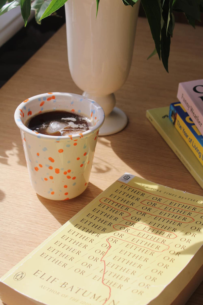 Naro Ceramic Cup in cream with orange and blue speckled dot pattern, filled with iced black coffee on a wooden surface with a book and white vase in natural sunlight.