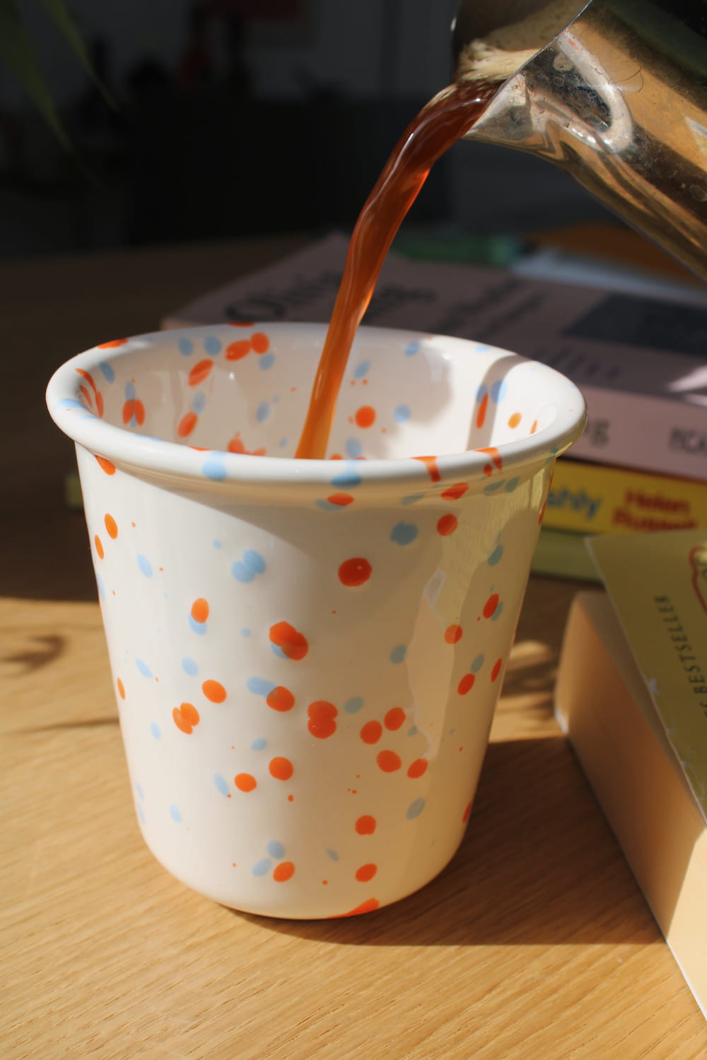 Naro Ceramic Cup in cream with orange and blue speckled dot pattern, filled with iced black coffee on a wooden surface with a book and white vase in natural sunlight.