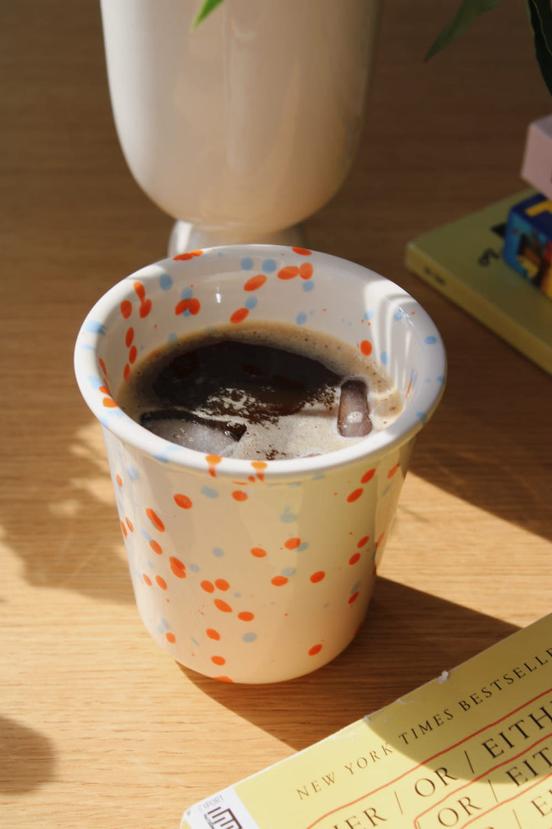 Naro Ceramic Cup in cream with orange and blue speckled dot pattern, filled with iced black coffee on a wooden surface with a book and white vase in natural sunlight.
