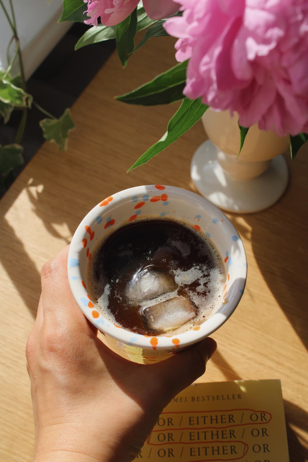 Naro Ceramic Cup in cream with orange and blue speckled dot pattern, filled with iced black coffee on a wooden surface with a book and white vase in natural sunlight.