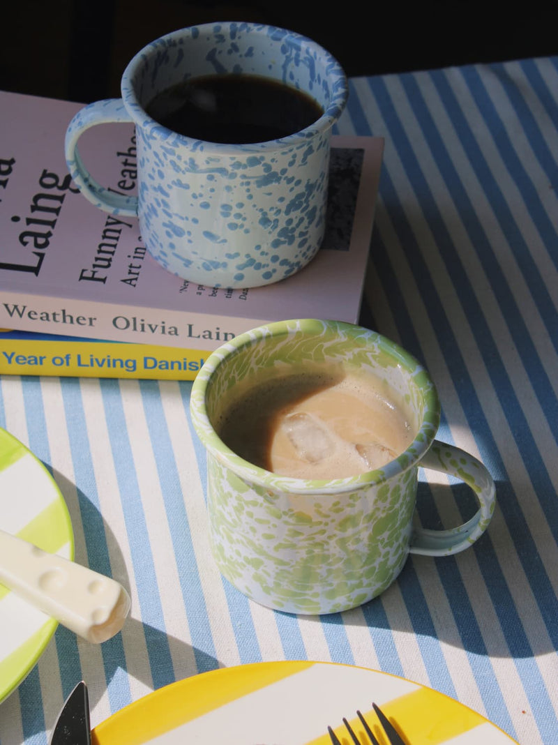Aeris Enamel Mug in blue speckle with black coffee and Clover Enamel Mug in green speckle with iced coffee, styled together on a blue striped surface with stacked books in natural sunlight.