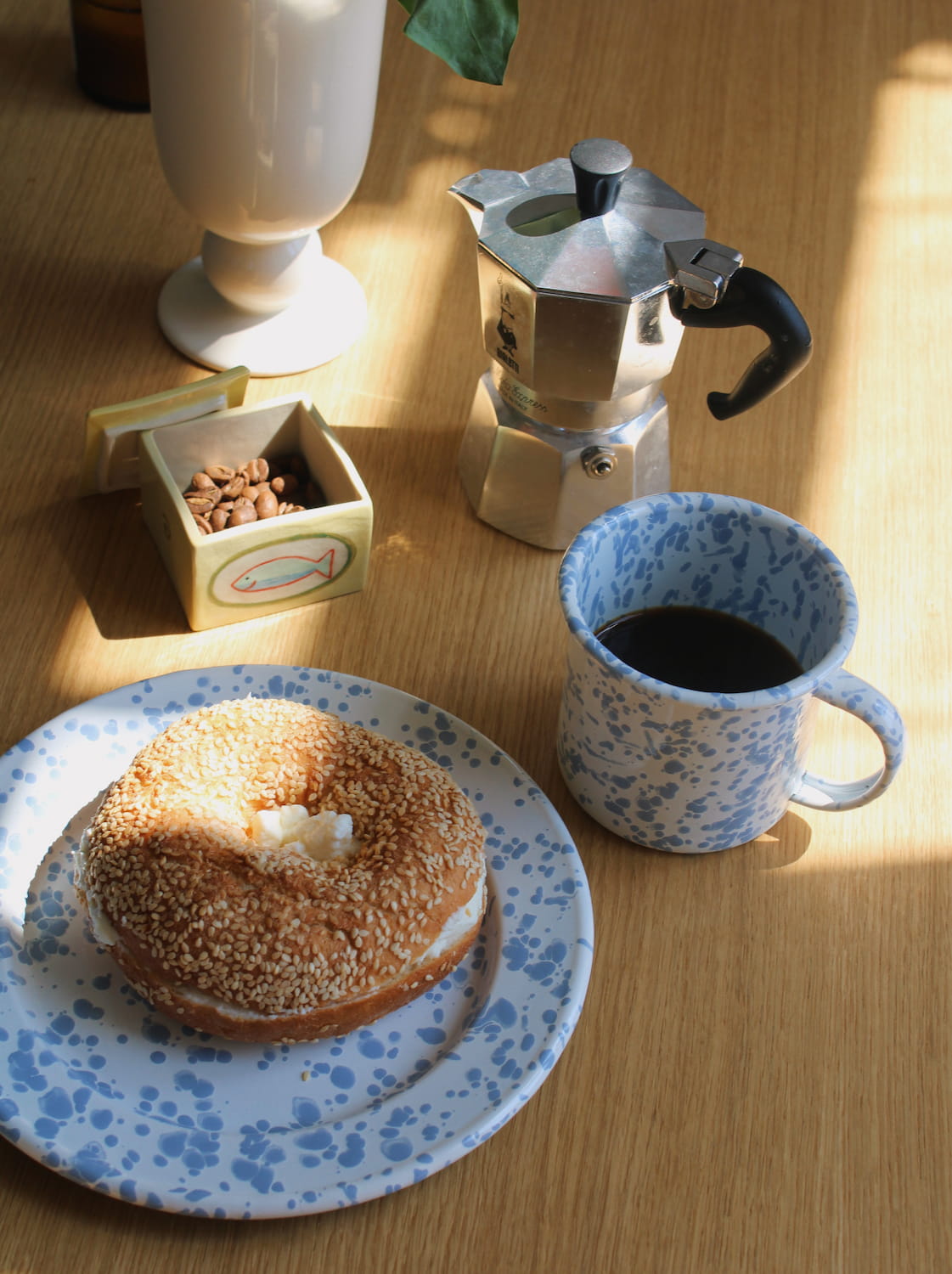 Aeris Enamel Mug in blue speckle filled with black coffee alongside the Aeris Enamel Plate with a sesame bagel and wooden cutlery, on a brown gingham surface in natural sunlight.