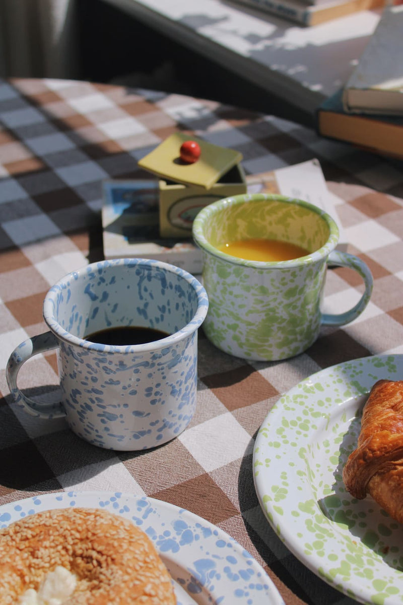 Aeris Enamel Mug in blue speckle with black coffee and Clover Enamel Mug in green speckle with iced coffee, styled together on a blue striped surface with stacked books in natural sunlight.