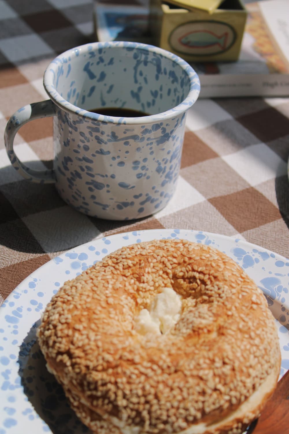 Aeris Enamel Mug in blue speckle filled with black coffee alongside the Aeris Enamel Plate with a sesame bagel and wooden cutlery, on a brown gingham surface in natural sunlight.