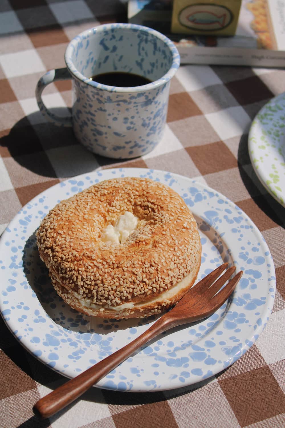 Aeris Enamel Mug in blue speckle filled with black coffee alongside the Aeris Enamel Plate with a sesame bagel and wooden cutlery, on a brown gingham surface in natural sunlight.