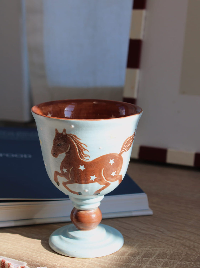 Fable Handmade Ceramic Horse Goblet in pale blue with hand-painted galloping horse and star detail, held in hand with lit candles in the blurred background in warm natural light.