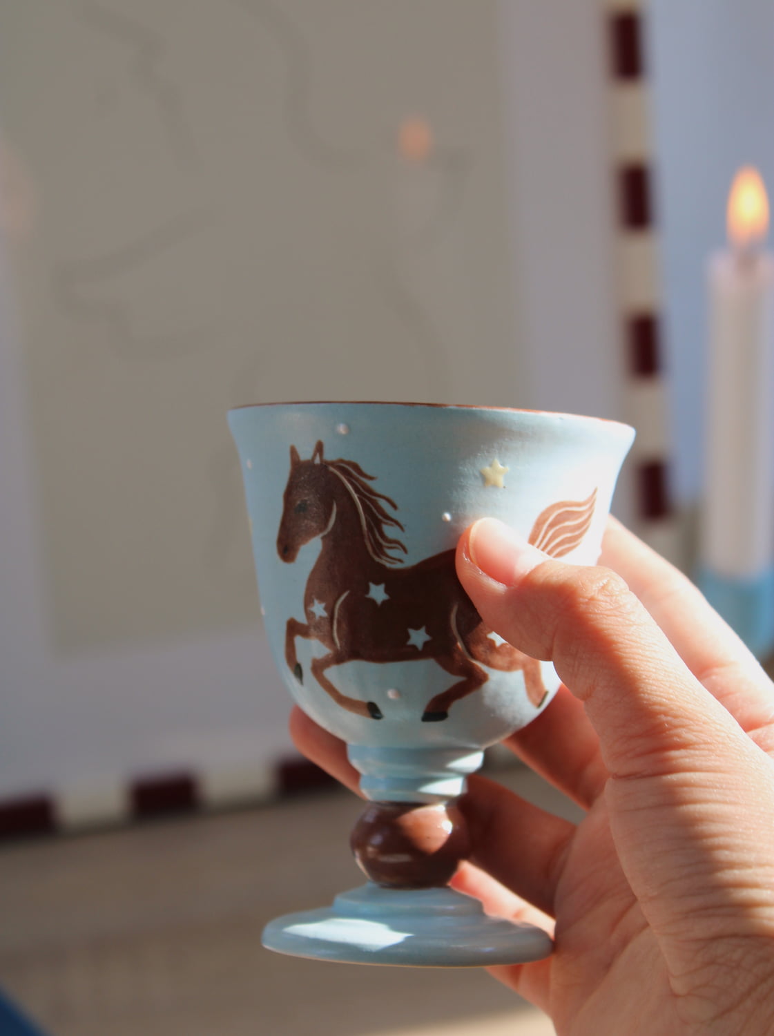 Fable Handmade Ceramic Horse Goblet in pale blue with hand-painted galloping horse and star detail, held in hand with lit candles in the blurred background in warm natural light.
