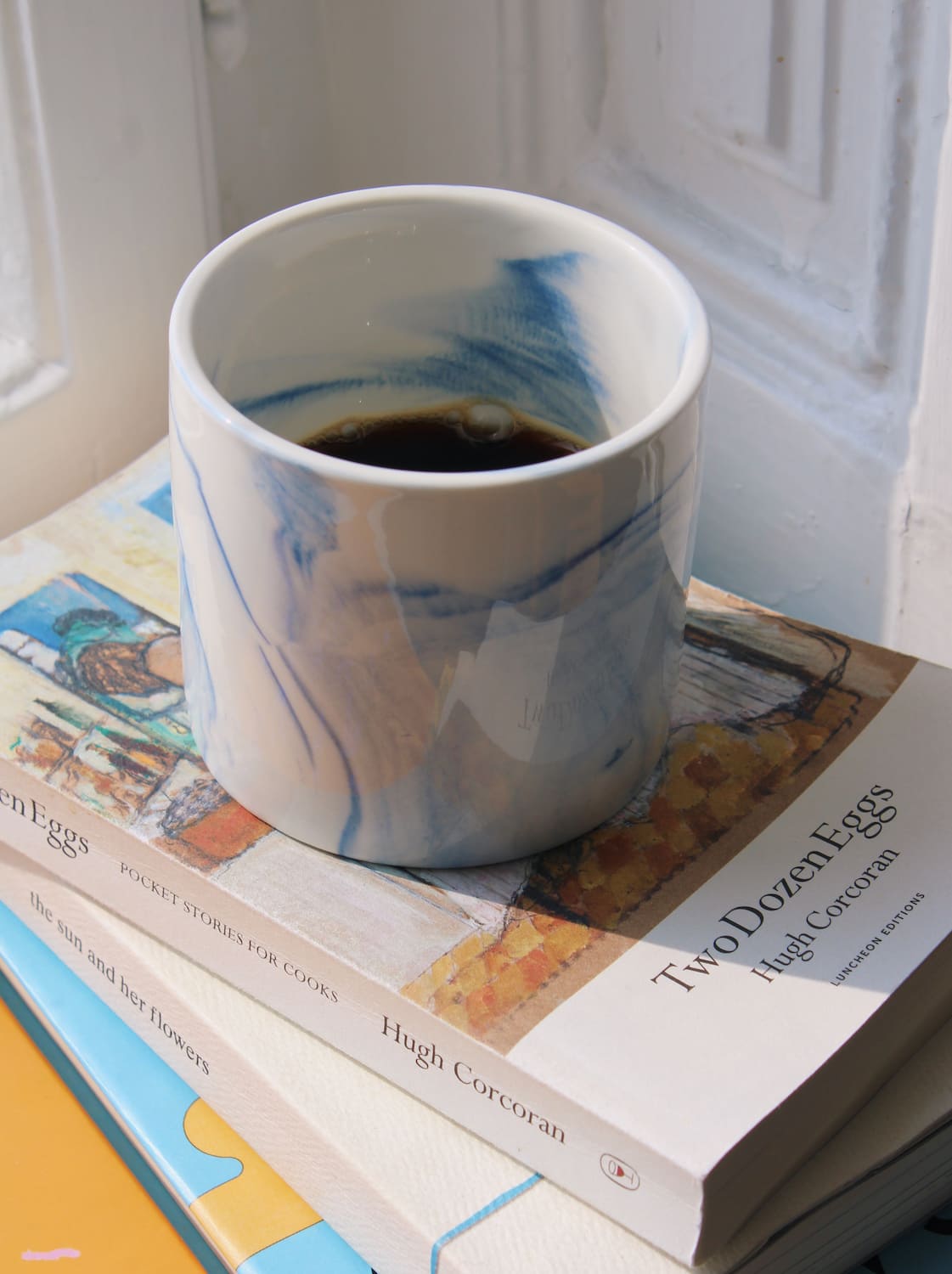 Sumi ceramic cup with soft blue brushstroke glaze placed on books by window in natural light.