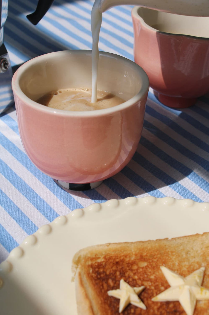 A pink ceramic cup filled with a brown beverage, with milk being poured into it, set on a blue and white striped tablecloth next to a plate of toast with star-shaped butter.