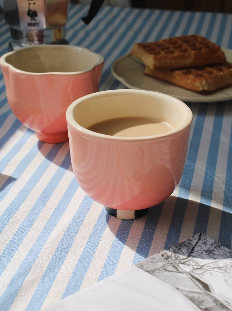 Two pink ceramic cups with scalloped rims sit on a blue and white striped tablecloth. The front cup is filled with a light-colored beverage, and a plate with waffles is blurred in the background.