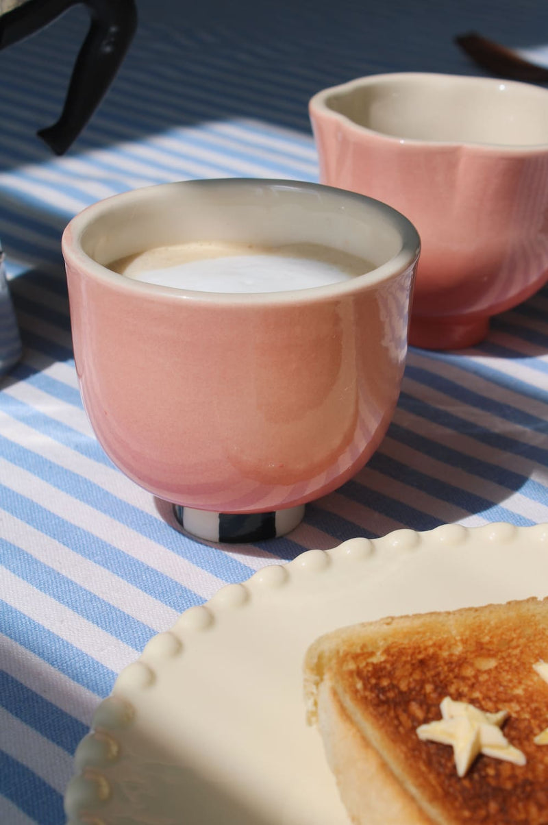 A pink ceramic cup filled with a light-colored beverage sits on a blue-and-white striped tablecloth next to a plate with toast. A second matching pink cup is blurred in the background.