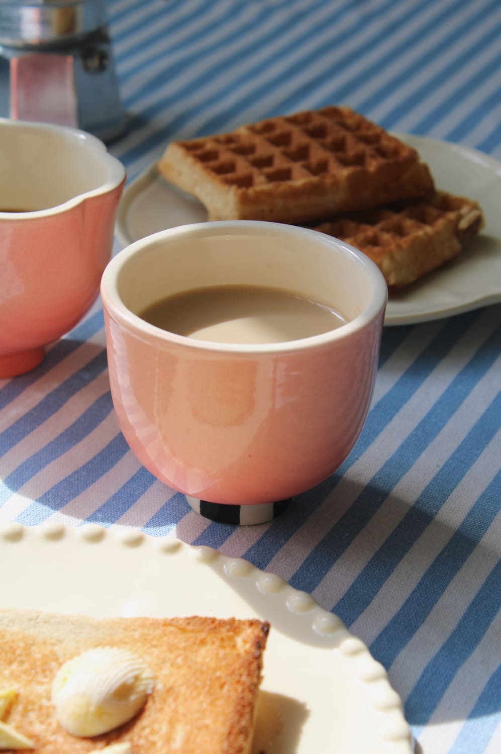 A pink ceramic cup filled with a light brown beverage sits on a blue-and-white striped tablecloth beside waffles and toast on white plates.
