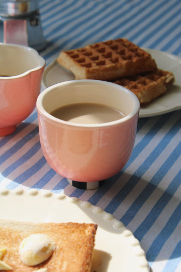 A pink ceramic cup filled with a light brown beverage sits on a blue-and-white striped tablecloth beside waffles and toast on white plates.