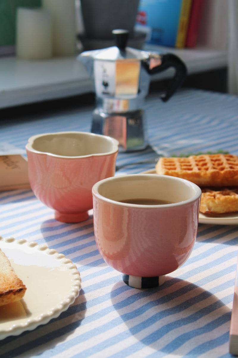 Two pink ceramic cups with white interiors and small black feet, set on a blue-and-white striped tablecloth with waffles, toast, and a moka pot in the background.