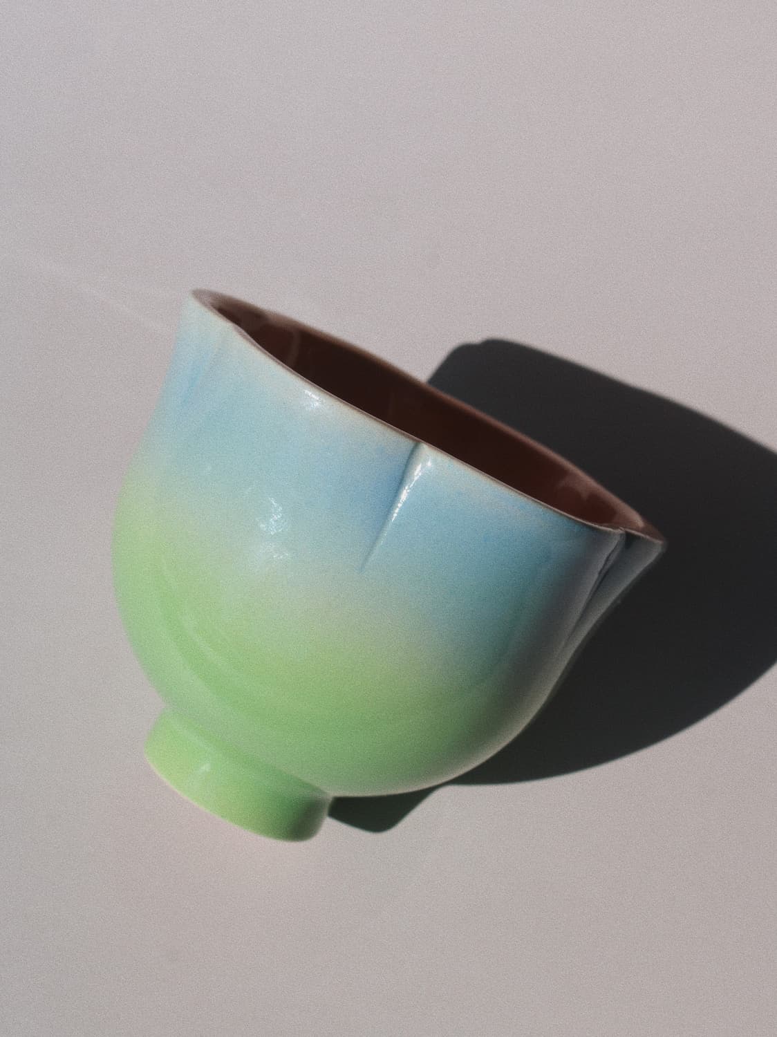 A ceramic bowl with a smooth gradient glaze transitioning from light blue at the top to pale green at the bottom, with a dark brown interior, photographed on a light gray background with a sharp shadow.
