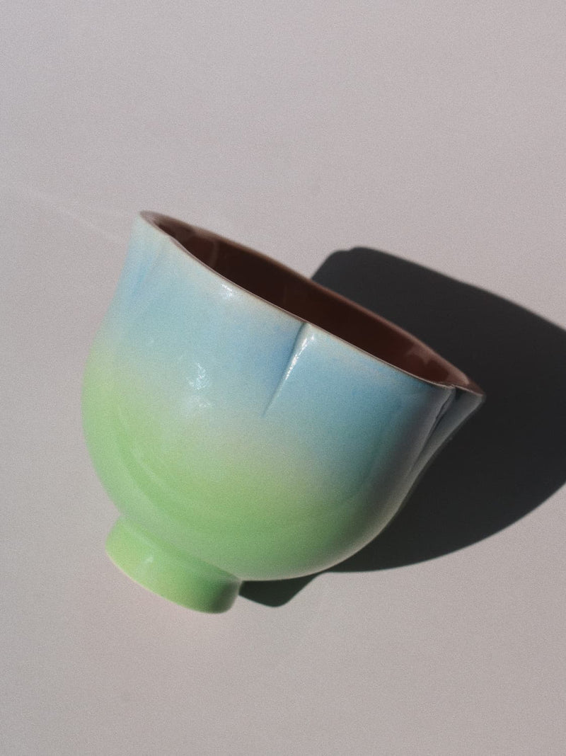 A ceramic bowl with a smooth gradient glaze transitioning from light blue at the top to pale green at the bottom, with a dark brown interior, photographed on a light gray background with a sharp shadow.