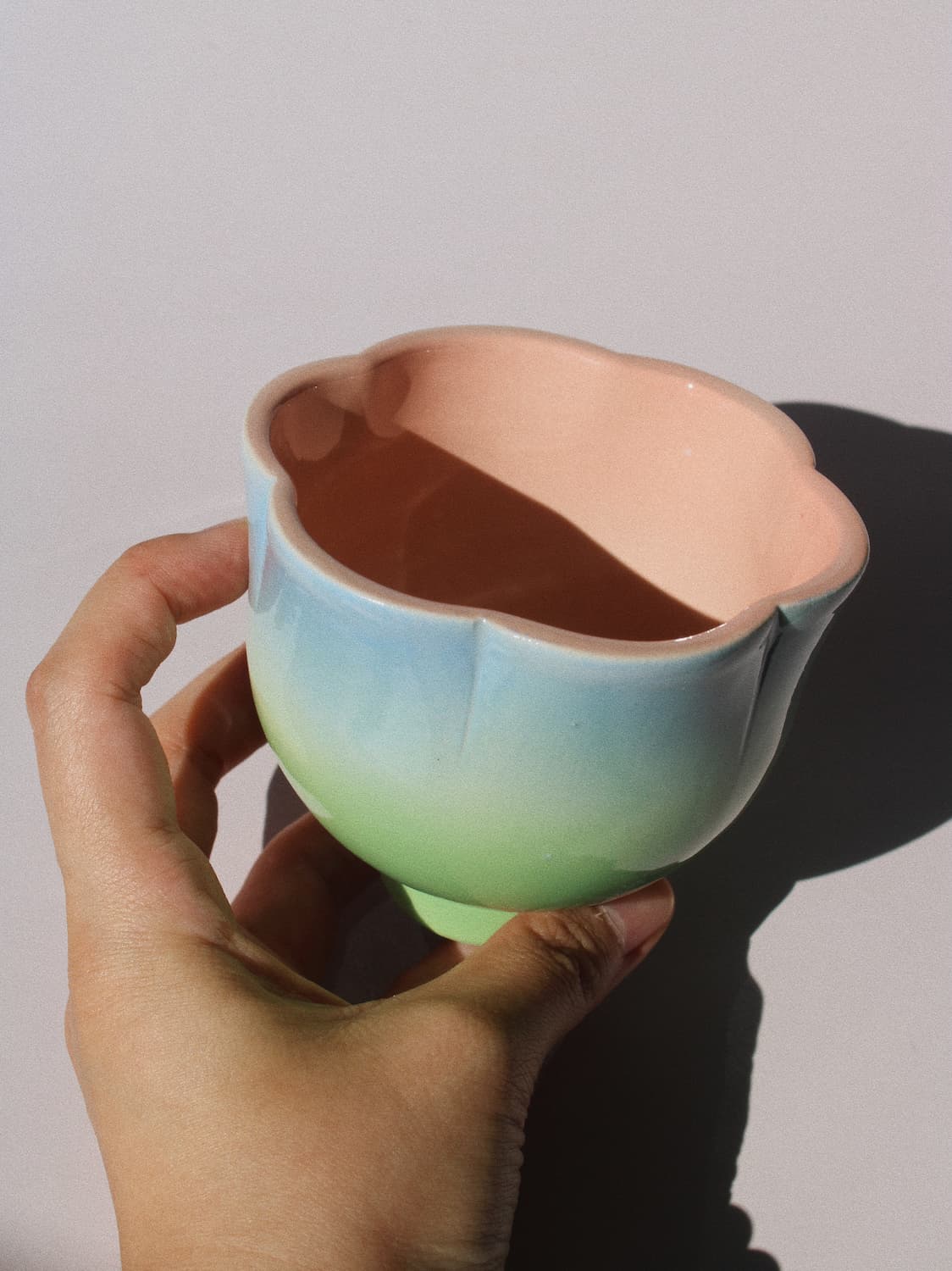 A hand holding a small ceramic cup with a pastel gradient from light blue to mint green on the outside and a peach-pink interior, set against a plain light gray background.