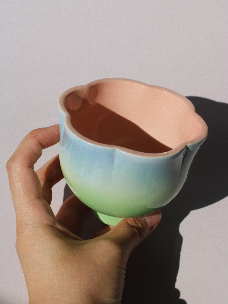 A hand holding a small ceramic cup with a pastel gradient from light blue to mint green on the outside and a peach-pink interior, set against a plain light gray background.