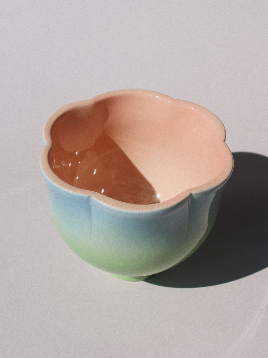 A ceramic flower-shaped bowl with a smooth pastel gradient glaze, transitioning from light blue at the base to soft green and peach-pink at the rim, shown on a neutral light gray background with a sharp shadow.