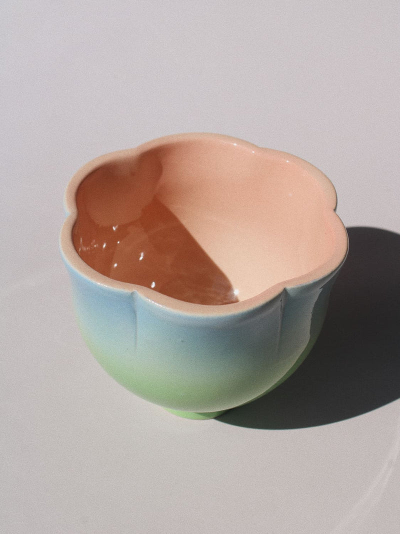 A ceramic flower-shaped bowl with a smooth pastel gradient glaze, transitioning from light blue at the base to soft green and peach-pink at the rim, shown on a neutral light gray background with a sharp shadow.